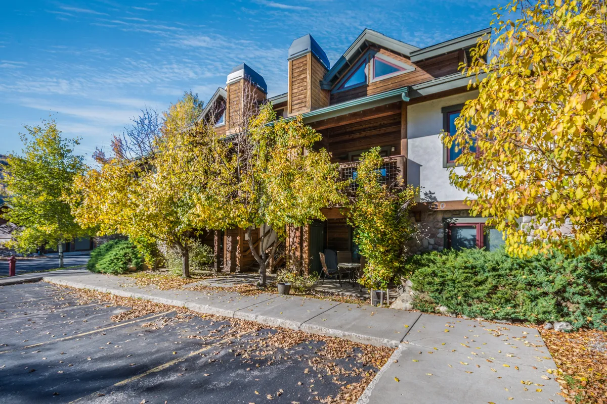 An exterior scene with colorful autumn trees, the building's stone and wood architecture, and front porch seating. The image evokes a sense of community gathering and access to outdoor activities, with pathways suggesting walkable access to local shops and dining.