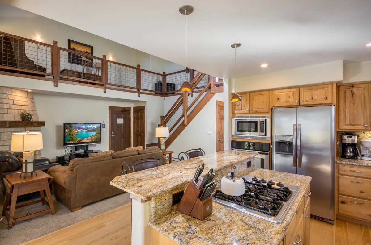 The image showcases a sun-filled, open-concept living area with soaring ceilings, a sleek granite kitchen island perfect for gathering, a modern open staircase, plush seating for relaxation, and a television by a cozy fireplace. The scene suggests warmth, comfort, and sociable spaces ideal for both entertaining and unwinding after a day on the slopes.