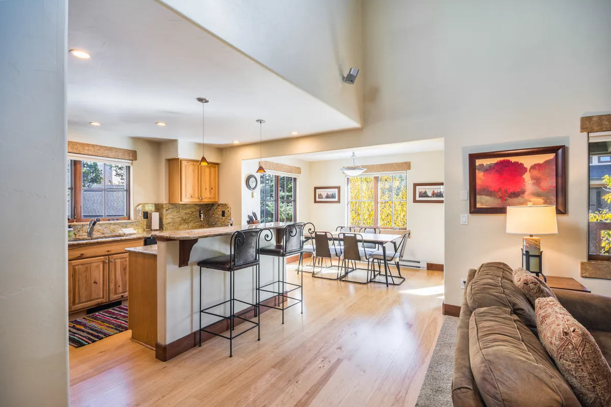 A view of the kitchen, dining, and living room arranged seamlessly together, with a kitchen island perfect for meal prep, a dining table for shared meals, a comfortable couch, and large windows letting in natural light. The room feels cozy and is set up for both relaxing and entertaining guests, with scenic views visible outside.