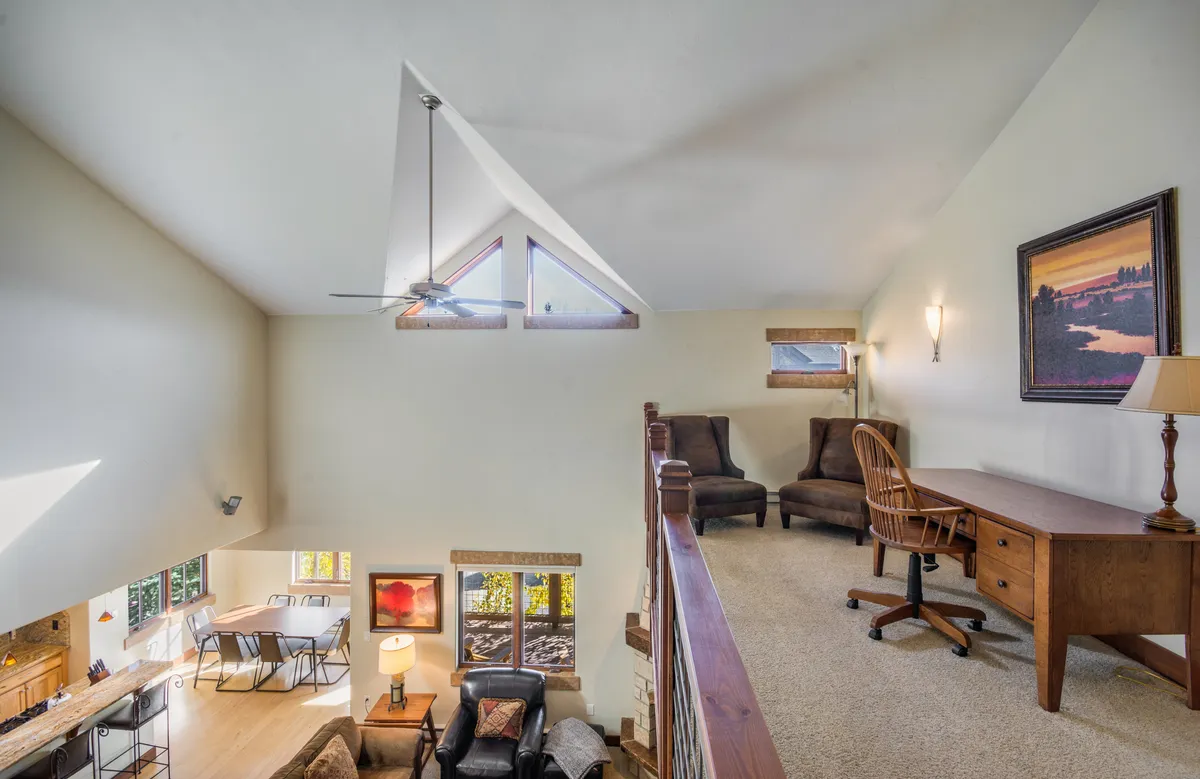 This image highlights a loft space featuring large windows that let in abundant natural light, a comfortable seating area, and a wooden desk. The setting appears calm and inviting, offering both a workspace and a cozy spot to relax or read, perfectly aligning with the idea of a versatile loft for work, play, and escape.