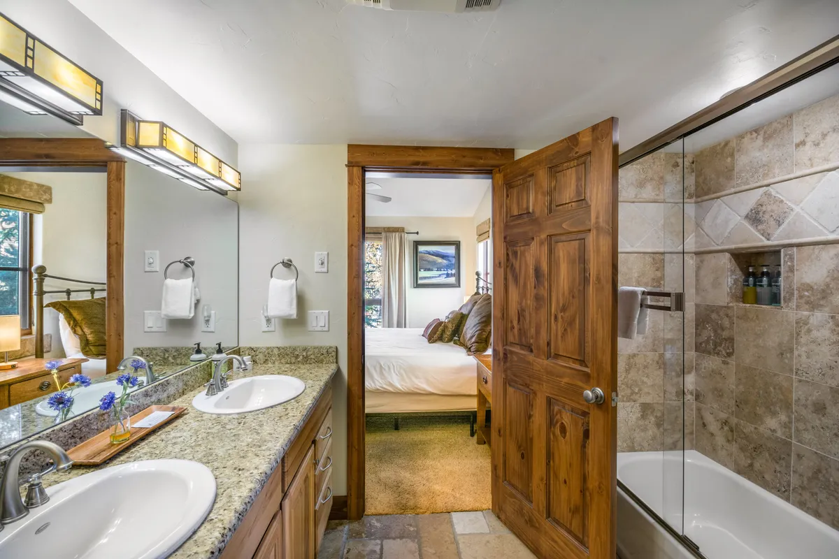 A tranquil bathroom with a double sink vanity, wooden door, shower area, and a glimpse of a comfortable bed in an adjacent room. The space exudes a spa-like atmosphere, perfect for unwinding and starting or ending the day refreshed.