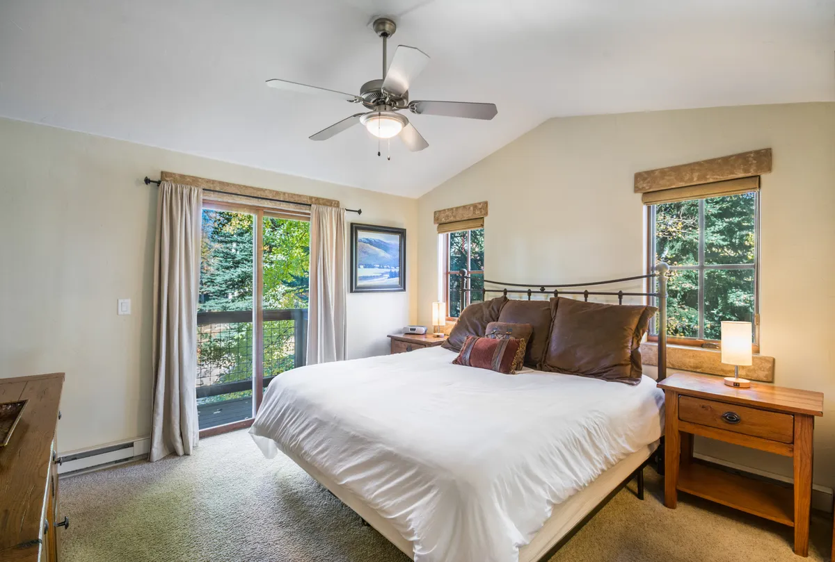 A serene bedroom featuring a comfortable bed set against large windows with drapes, accompanied by nightstands and artwork. Natural light fills the room, creating an inviting and restful atmosphere ideal for relaxation and recharging.
