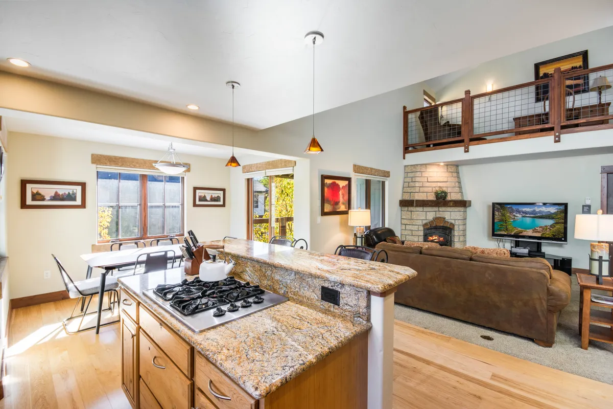 A spacious open-plan living area featuring a granite kitchen island with bar stools, a wooden dining table set for a meal, a cozy stone fireplace, and an open loft above. Warm lighting and ample natural light from large windows create a welcoming atmosphere. The space shows several people gathering for breakfast, relaxing by the fire, and celebrating special occasions.